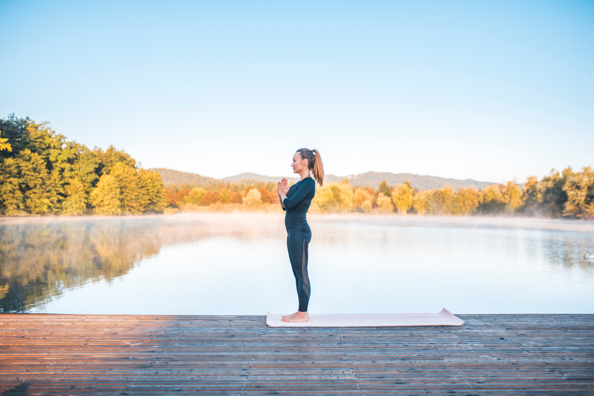 blonde woman with ponytail wearing all black standing on wooden dock in mountain yoga pose