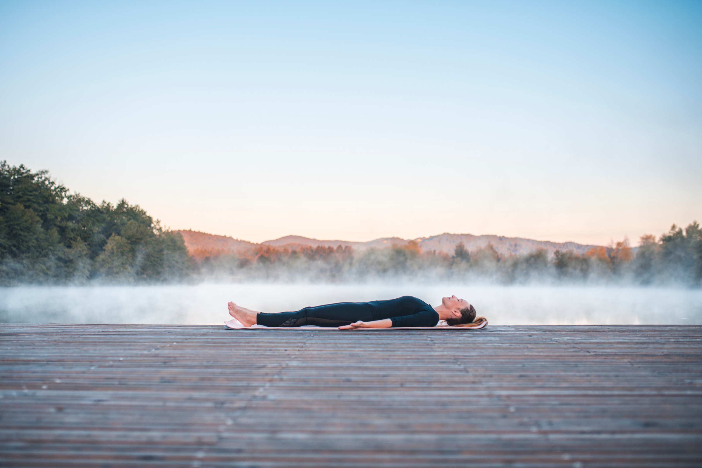 blonde woman with ponytail in all black on a wooden dock by water doing corpse svasana yoga pose