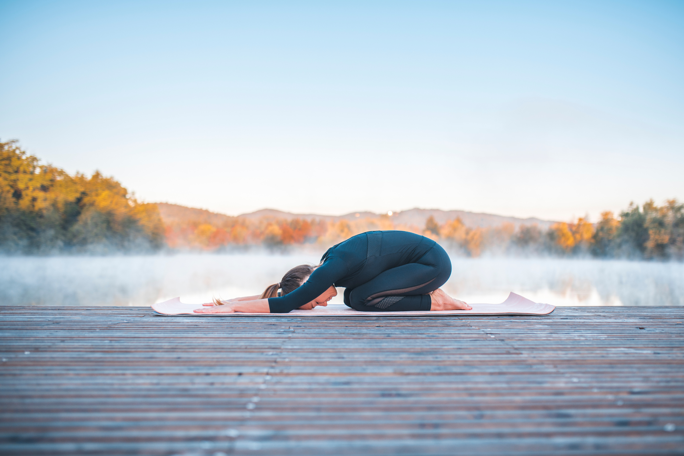 blonde woman with ponytail wearing all black on a wooden dock by water doing child pose yoga pose