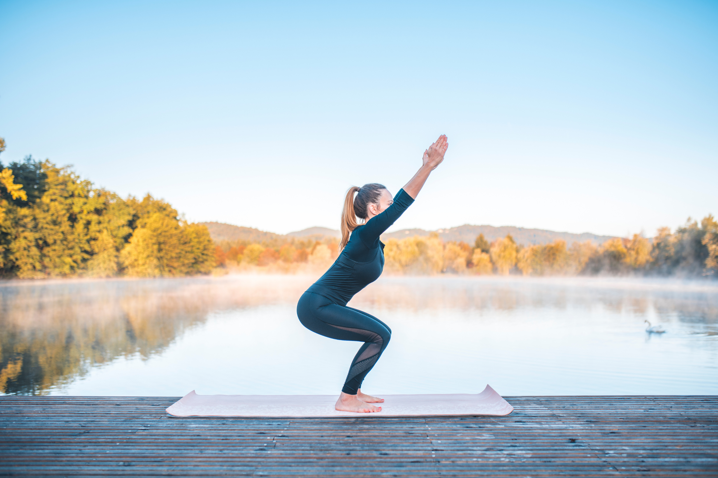 blonde woman with ponytail in all black on a wooden dock by water doing chair yoga pose