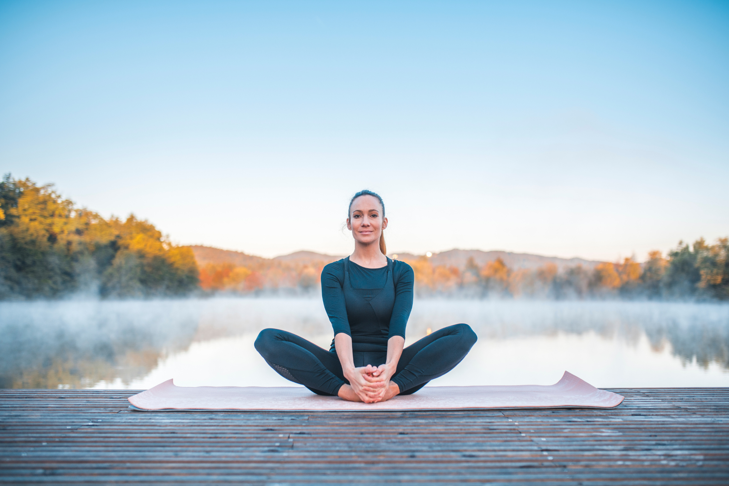 blonde woman with ponytail wearing all black on a wooden dock by water doing bound angle yoga pose
