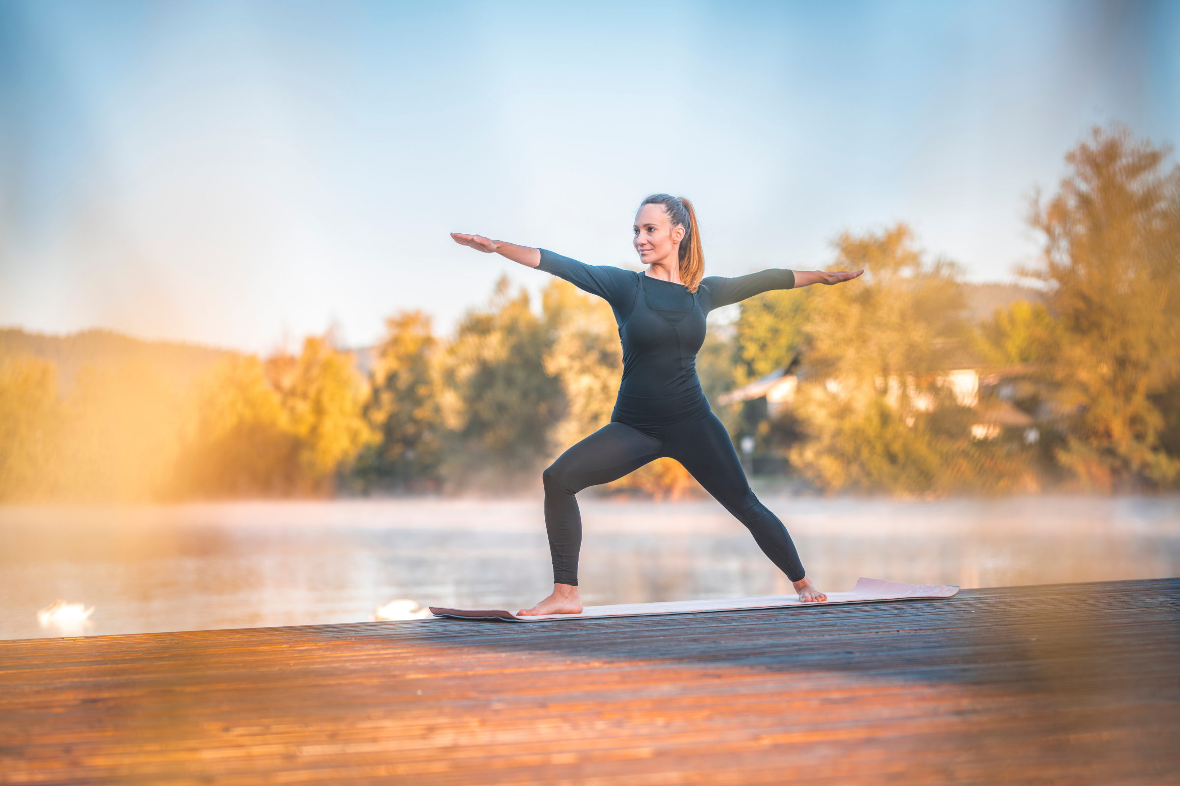 blonde woman with ponytail wearing all black on a wooden dock by water doing warrior II yoga pose