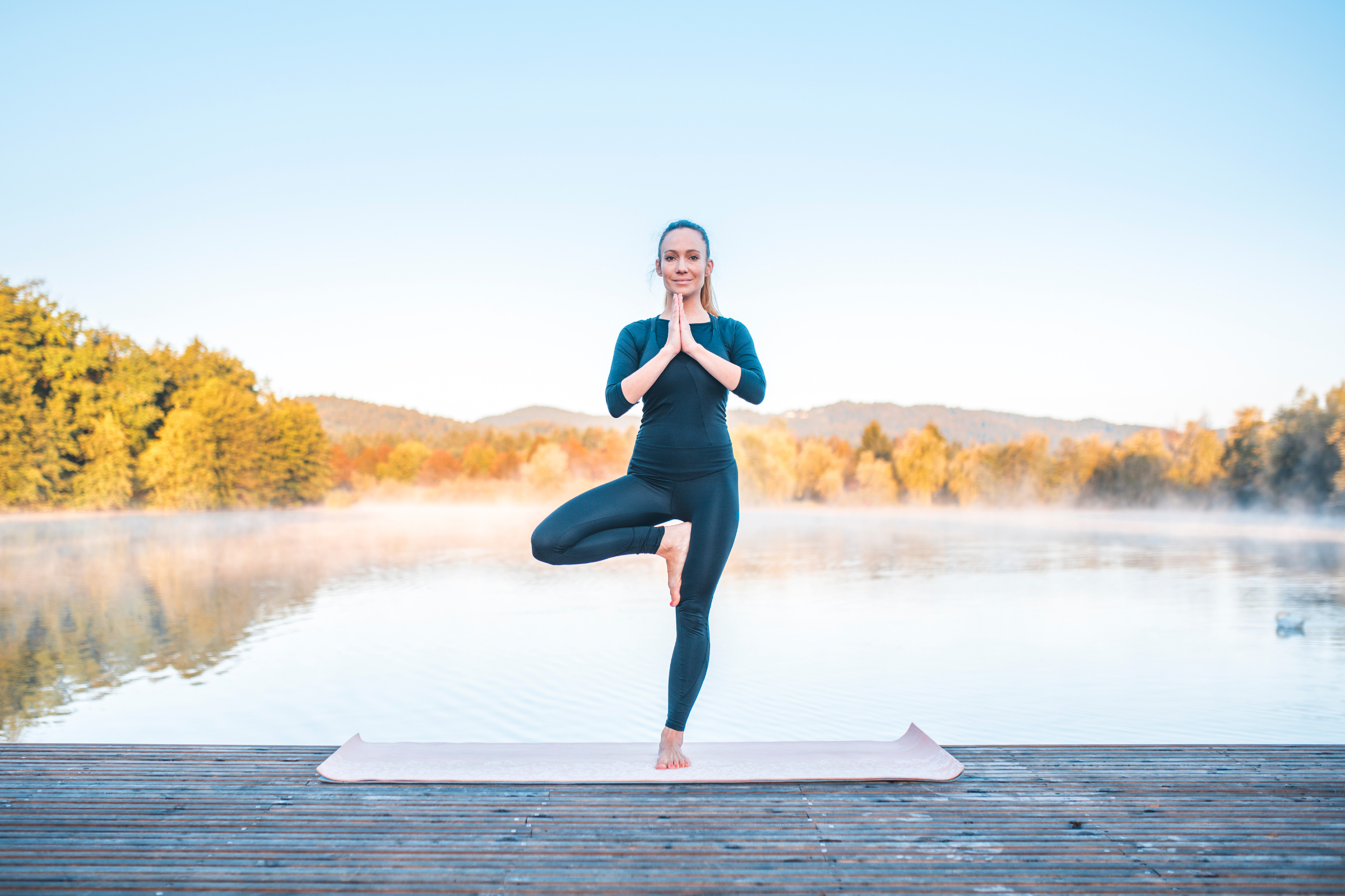 blonde woman with ponytail wearing all black standing on wooden dock in tree yoga pose