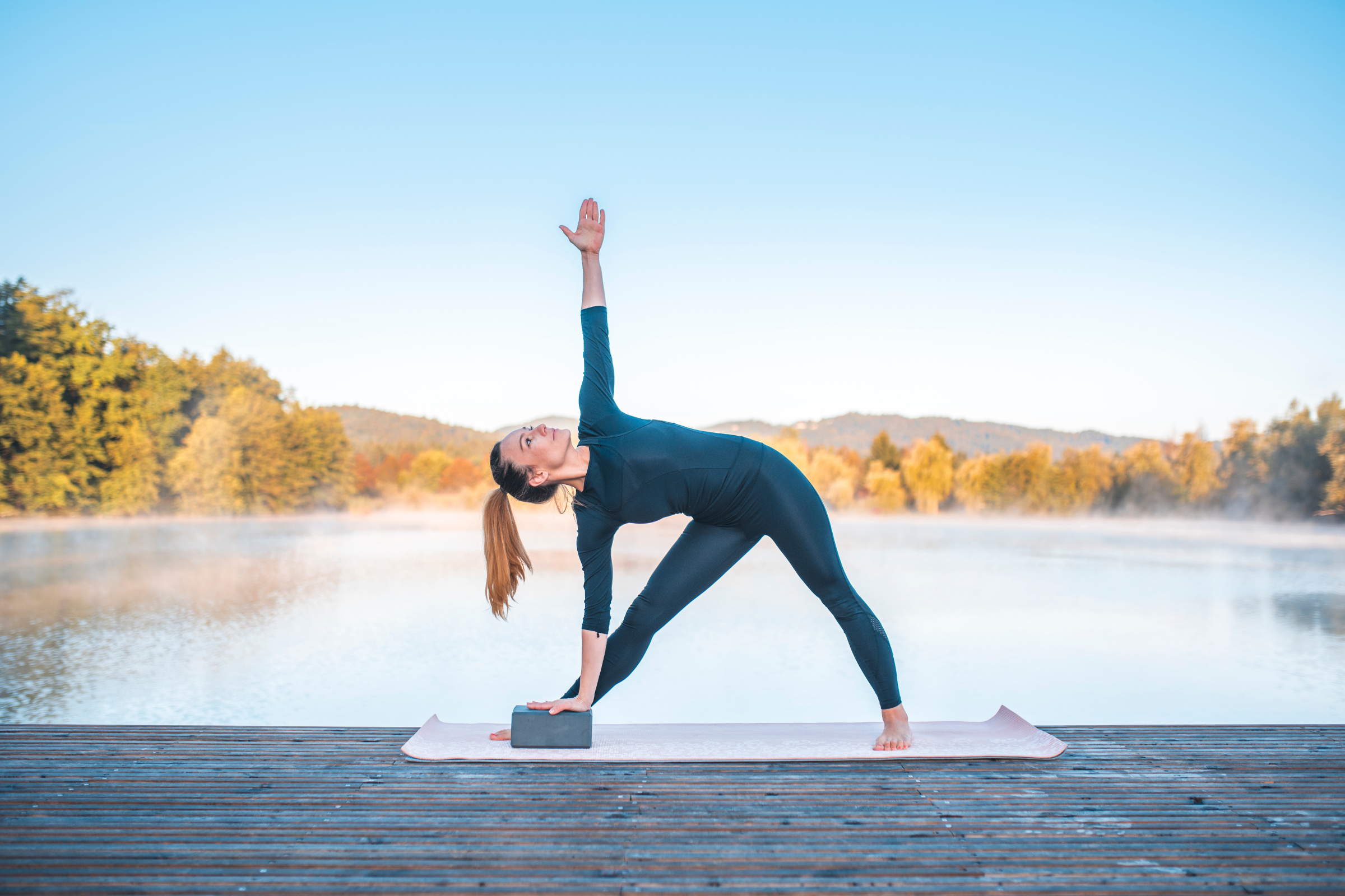 blonde woman with ponytail wearing all black on a wooden dock by water doing triangle yoga pose