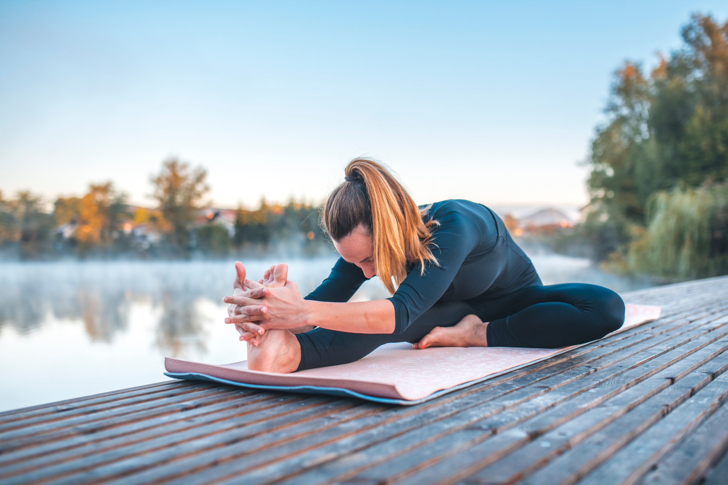 blonde woman with ponytail wearing all black on a wooden dock by water doing head to knee forward bend pose