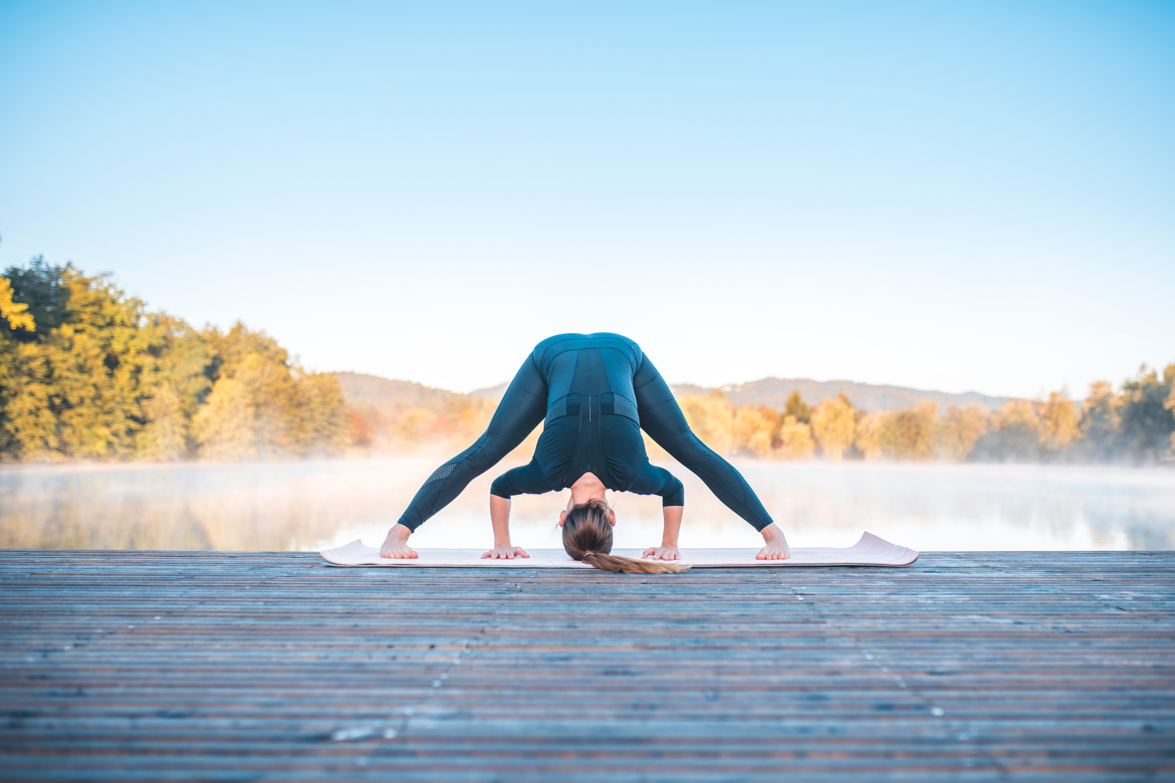 blonde woman with ponytail wearing all black on a wooden dock by water doing wide legged forward bend yoga pose